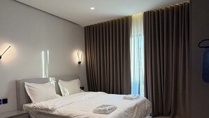 Et moderne hotelværelse med en stor seng, et sengebord, en lampe og udsigt til det fri gennem gardiner.