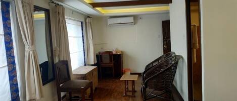 A room with a wooden floor, a desk, a chair, a small table, a wicker chair, and a window with curtains.