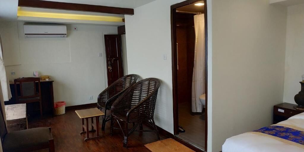 Quarto de hotel com uma cama, uma mesa pequena, duas cadeiras de vime, uma escrivaninha de madeira, ar condicionado e uma porta que dá para outro quarto.