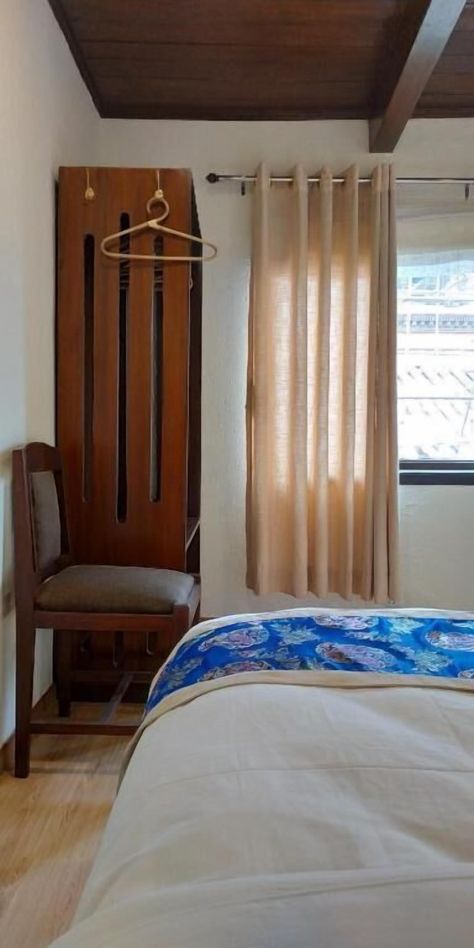 A bedroom with a bed, a chair, a wooden wardrobe, and a window with curtains.