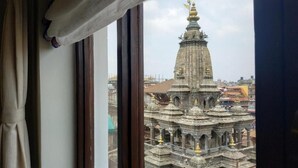 Une vue par la fenêtre sur un temple à l’architecture complexe, un rideau qui recouvre partiellement la fenêtre, et un aperçu du paysage urbain à l’extérieur.