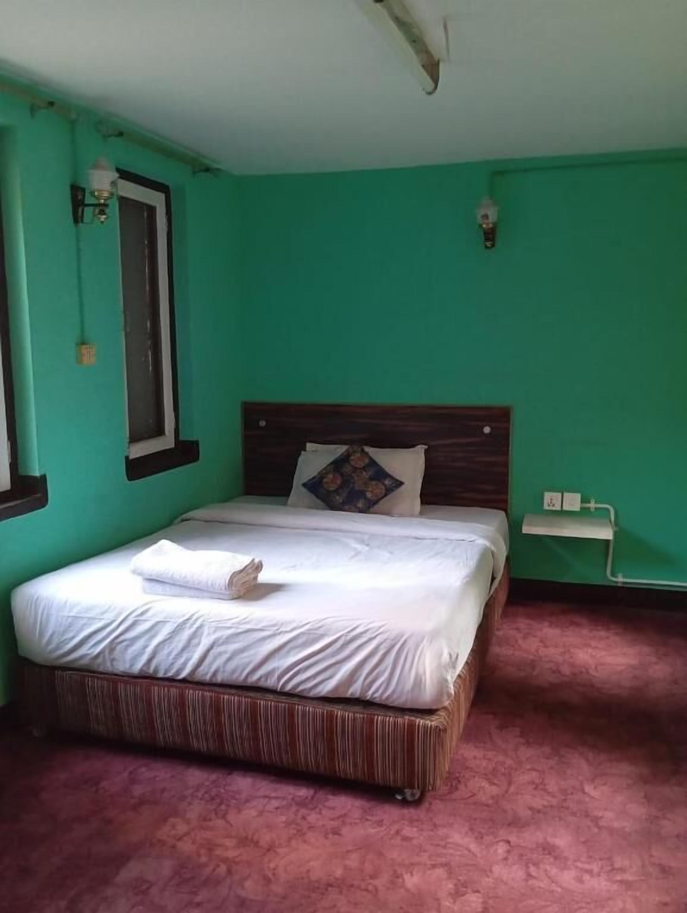A room with a bed, a window, a wall-mounted light fixture, and a small bedside table.