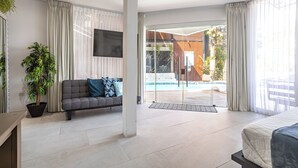 A modern hotel room with a sofa, a bed, a TV, a potted plant, and a view of a pool.