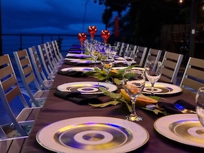 A long dining table set for a meal with chairs on both sides, a view of the sea in the background, and a dark evening sky. - Penthouse with Panoramic Ocean Views - Hotel VOS (Manuel Antonio)