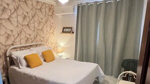 A bedroom with a bed, a desk, a chair, a ceiling fan, and a window with curtains. - The Distrito Atelier (Santo Domingo)