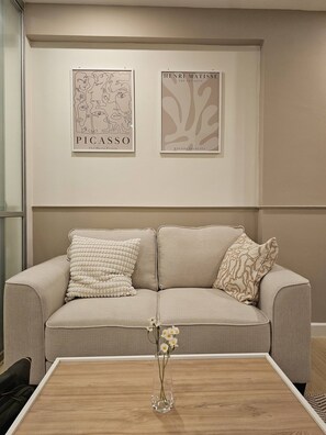 Ein beiges Sofa mit gemusterten Kissen, eine Glasvase mit Blumen und zwei gerahmte Kunstwerke an der Wand.