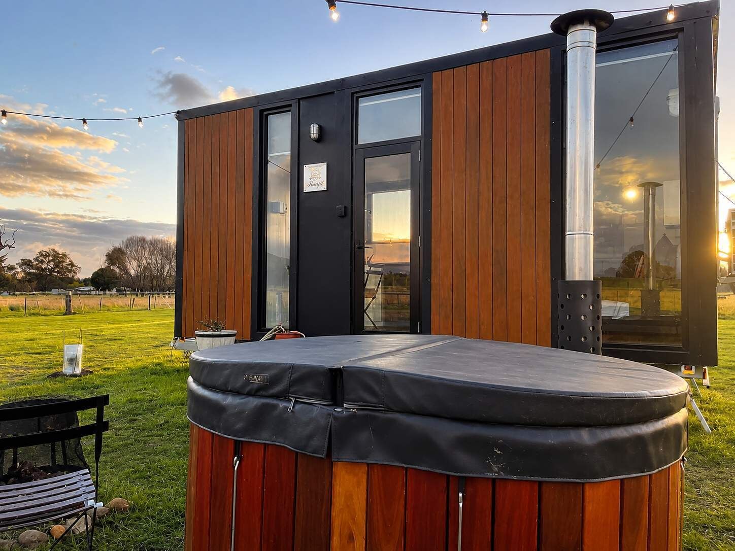A modern outdoor hot tub with a wooden exterior, a glass door, and a string of lights.