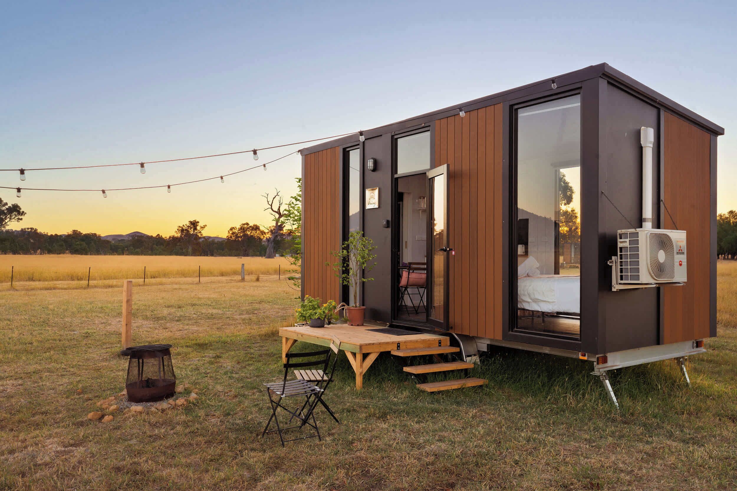 Une petite cabane moderne dotée d’une porte coulissante, d’une petite table avec des chaises, d’une plante en pot et d’un appareil de climatisation mobile.