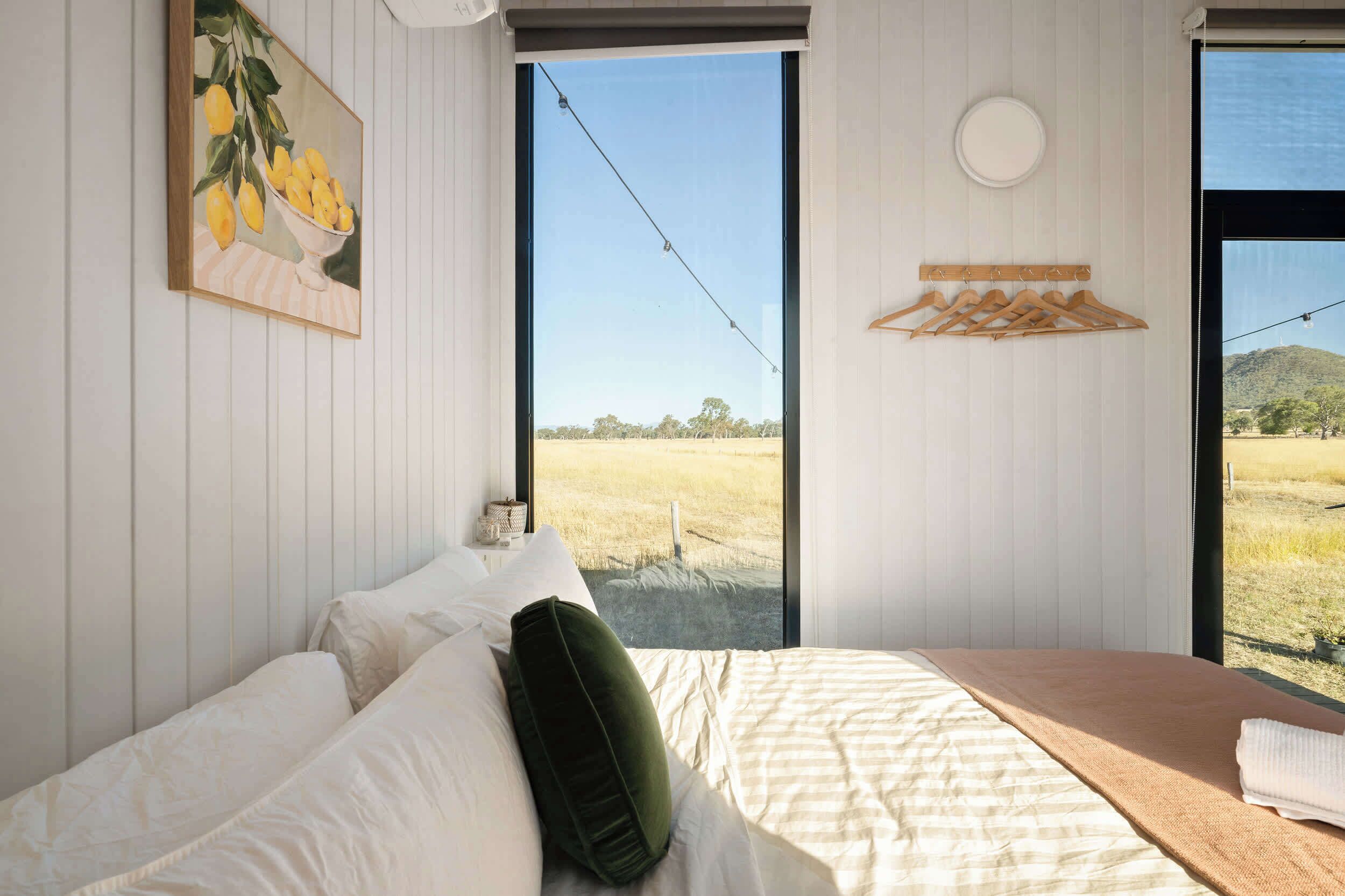 Un dormitorio con cama, almohadas, vista a un campo a través de un ventanal grande, un cuadro en la pared y un perchero de madera.