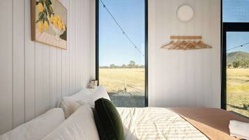 Un dormitorio con cama, almohadas, vista a un campo a través de un ventanal grande, un cuadro en la pared y un perchero de madera.