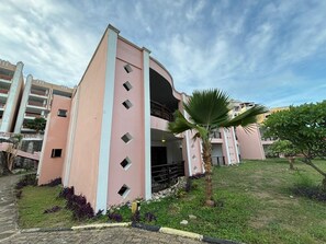 Un bâtiment rose avec une entrée centrale, entouré d’une pelouse et d’un palmier.