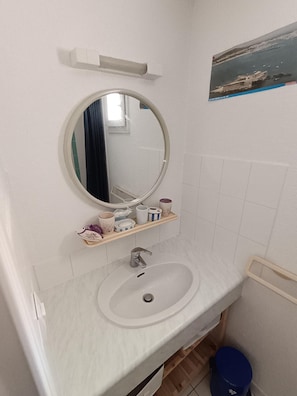 Ein Badezimmer mit rundem Spiegel, Waschbecken, Ablage mit Pflegeprodukten und blauem Mülleimer.