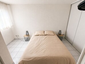 Ein kleines, minimalistisches Schlafzimmer mit einem Bett, zwei Nachttischen mit Lampen und einem Fenster mit transparenten Vorhängen.