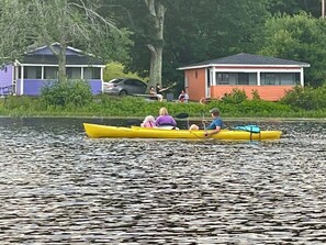 A yellow canoe on calm water with people paddling, surrounded by greenery and buildings in the background. - Cozy Waterfront Cottage Rental on the Shores of Harvey Lake in Northwood, New Hampshire (Northwood)