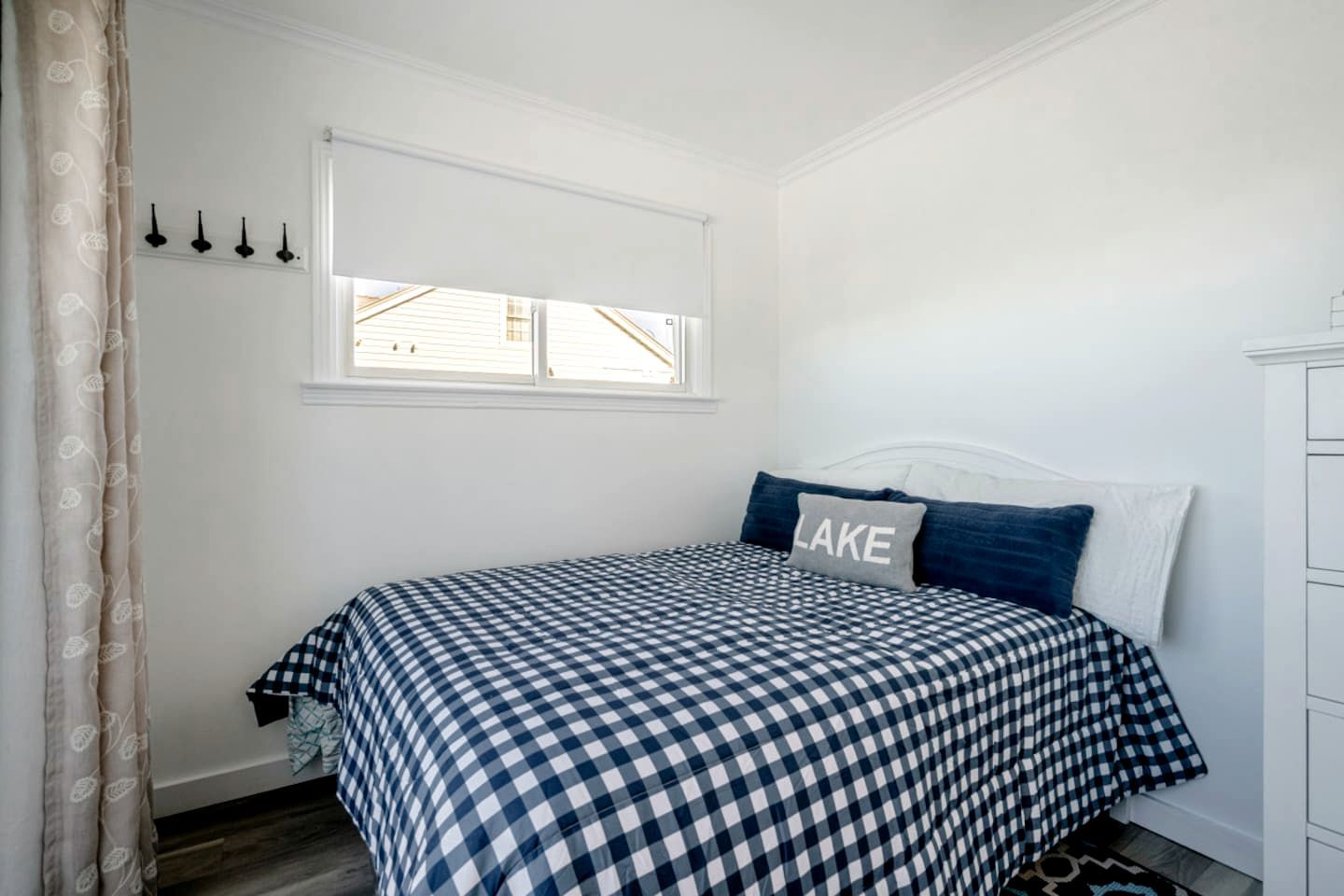 A bedroom with a checkered bedspread, a pillow with the word 'LAKE,' a window with blinds, and a white dresser.