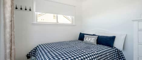 Une chambre avec une couvre-lit à carreaux, un coussin orné du mot «
LAKE
», une fenêtre avec des stores et une commode blanche.