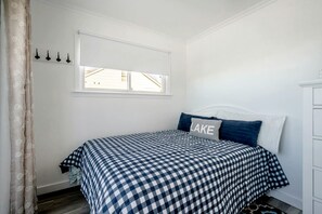 A bedroom with a checkered bedspread, a pillow with the word 'LAKE,' a window with blinds, and a white dresser. - Stunning Cottage with Nice Views and Fire Pit in Leamington, Ontario (Leamington)