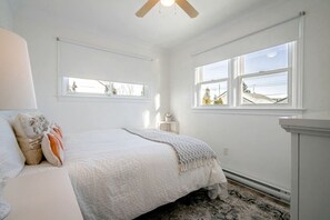 Quarto com cama, duas janelas, ventoinha no teto e radiador.