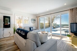 A modern living room with a sofa, TV, and large windows overlooking a snowy outdoor area. - Stunning Cottage with Nice Views and Fire Pit in Leamington, Ontario (Leamington)