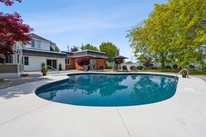 Une piscine résidentielle à bord incurvé, un patio couvert avec des chaises, et une maison en arrière-plan.