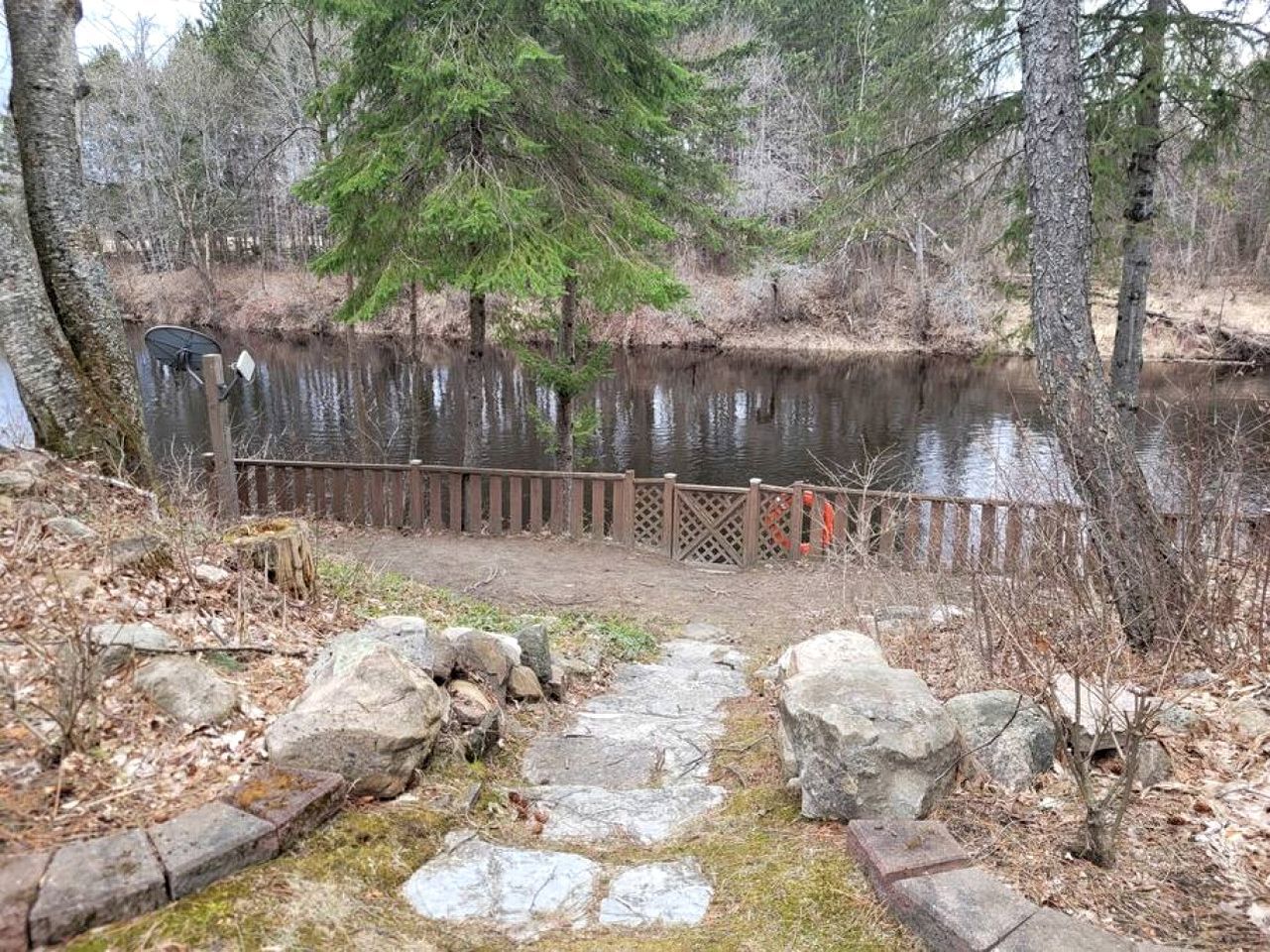 Ein Steinweg führt zu einem Holzzaun an einem Gewässer, umgeben von Bäumen und Felsen.