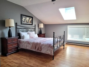 Une chambre à coucher comprenant un lit, deux tables de chevet, un luminaire au plafond, une fenêtre avec des stores et un puits de lumière.