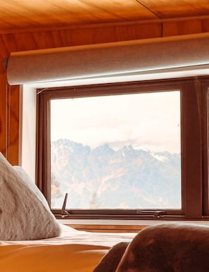 A room with a bed, a window with a view of mountains, and wooden walls.