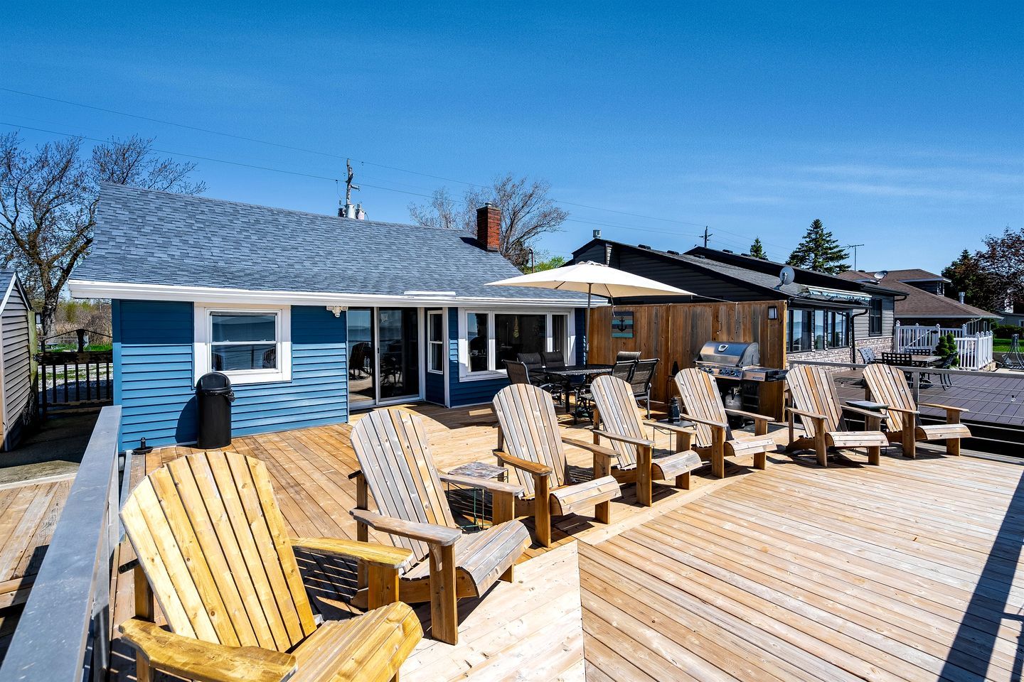 A deck with wooden chairs and a grill, a blue building with a covered patio, and a clear sky.