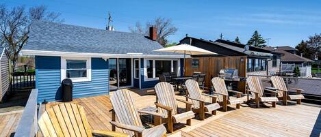 A deck with wooden chairs and a grill, a blue building with a covered patio, and a clear sky.