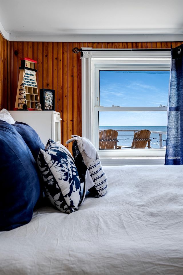 A bedroom with a large window, wooden paneling, a bed with patterned pillows, a small shelf with decorative items, and a view of the sea.