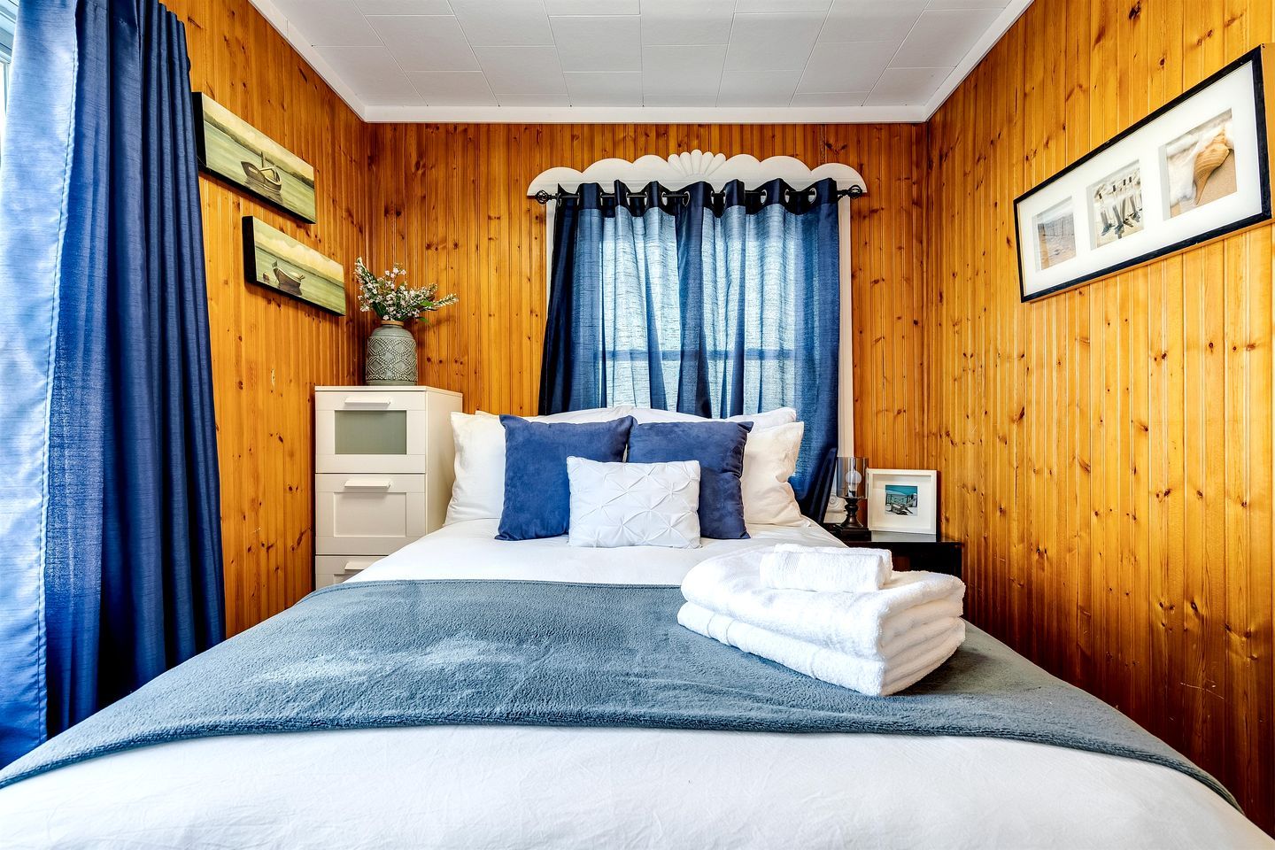 Une chambre avec un mur en bois, un lit avec une literie bleue, une table de chevet et une fenêtre avec des rideaux bleus.