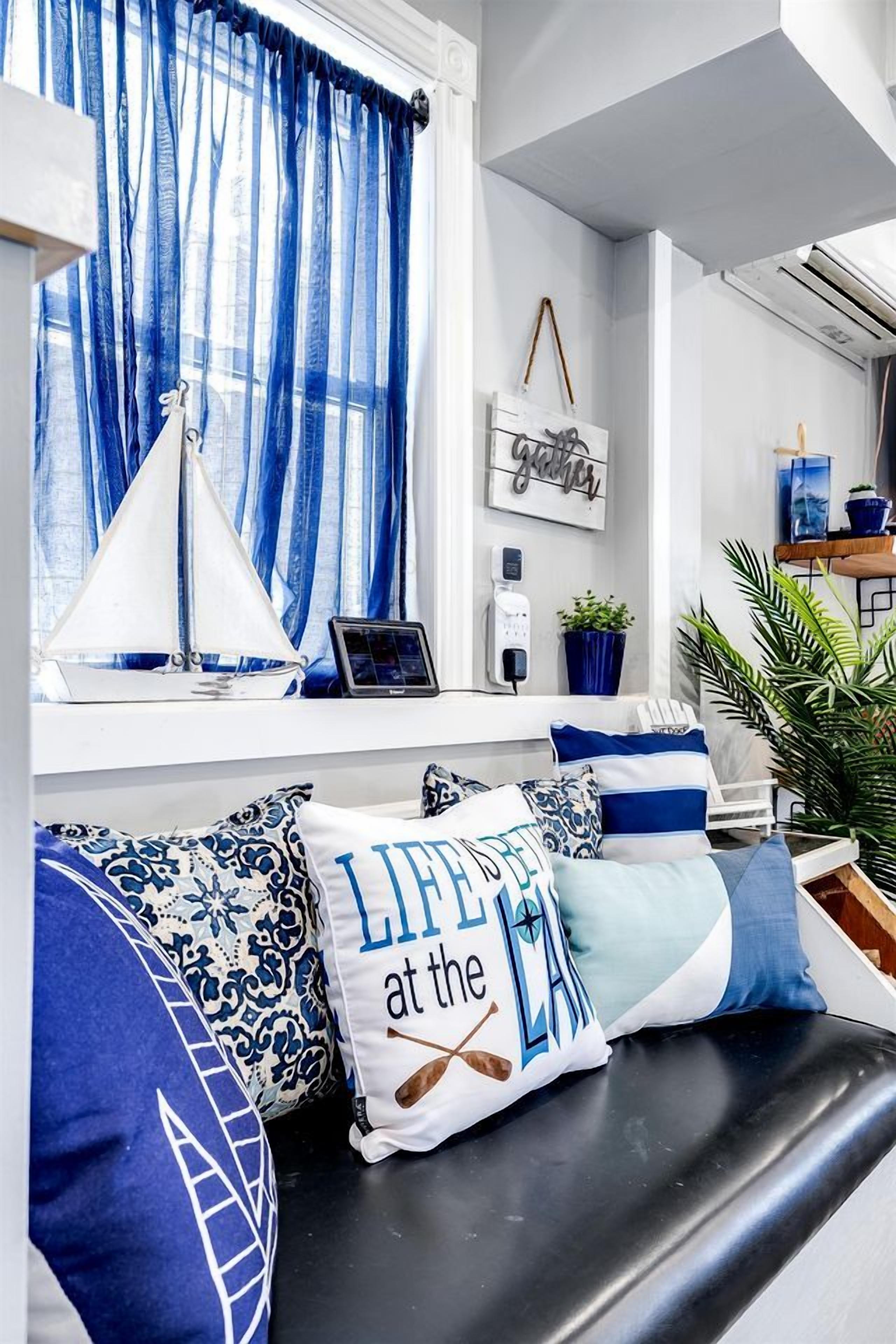 A modern living room with a black sofa, blue and white patterned cushions, a sailboat decoration, and a window with blue curtains.