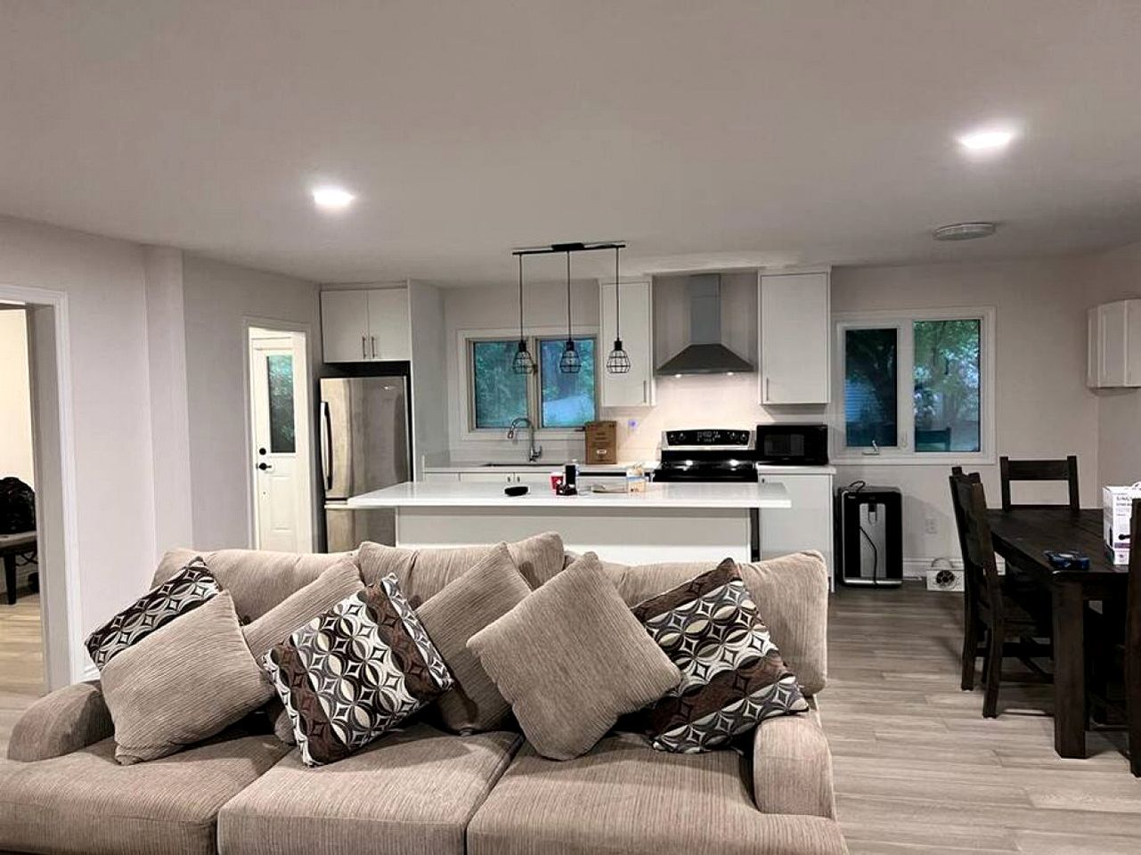 Una sala moderna con sofá, comedor con mesa y sillas, cocina con isla central, luces colgantes y vistas al exterior a través de ventanas.
