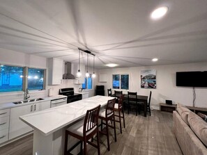 Une cuisine moderne avec un îlot central, un coin repas avec des chaises, un canapé et un téléviseur fixé au mur.