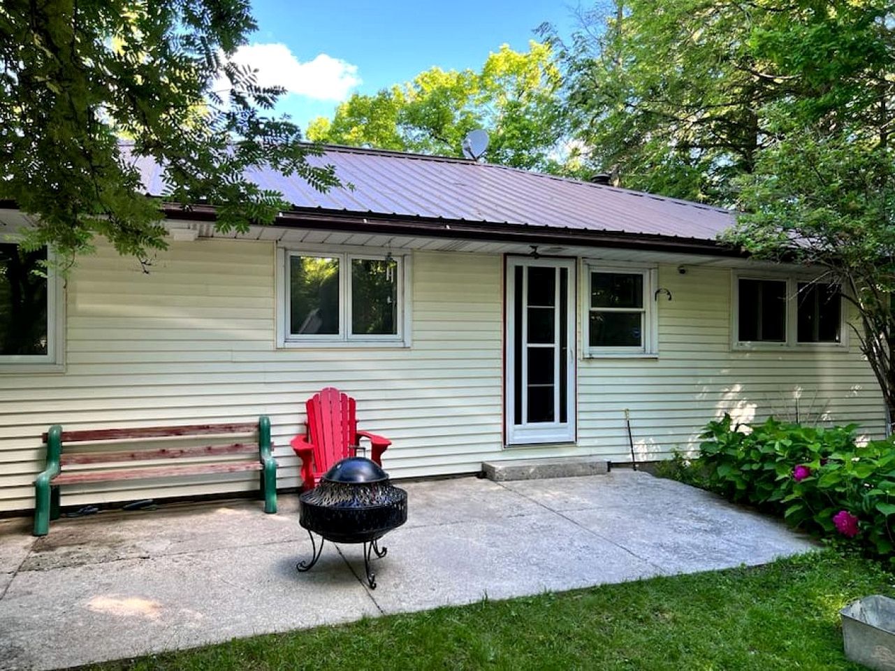 A single-story house with a metal roof, a front door, and a small porch with a bench and a red chair.