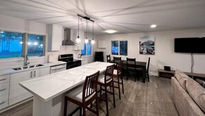 A modern kitchen with a central island, dining area with chairs, a sofa, and a TV mounted on the wall.