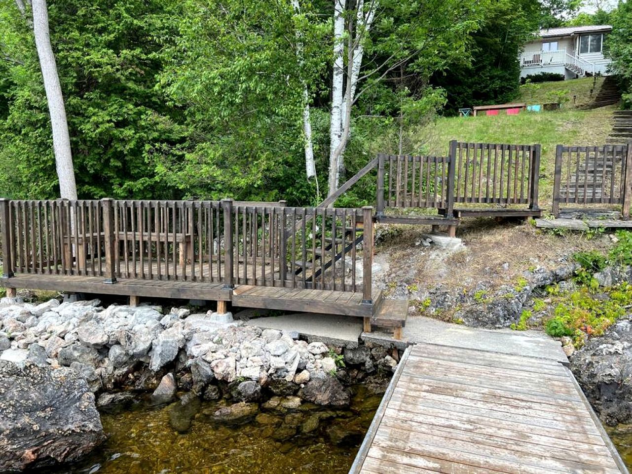 Un puente de madera sobre un arroyo pedregoso, con pasarela de madera y una zona delimitada por una valla.