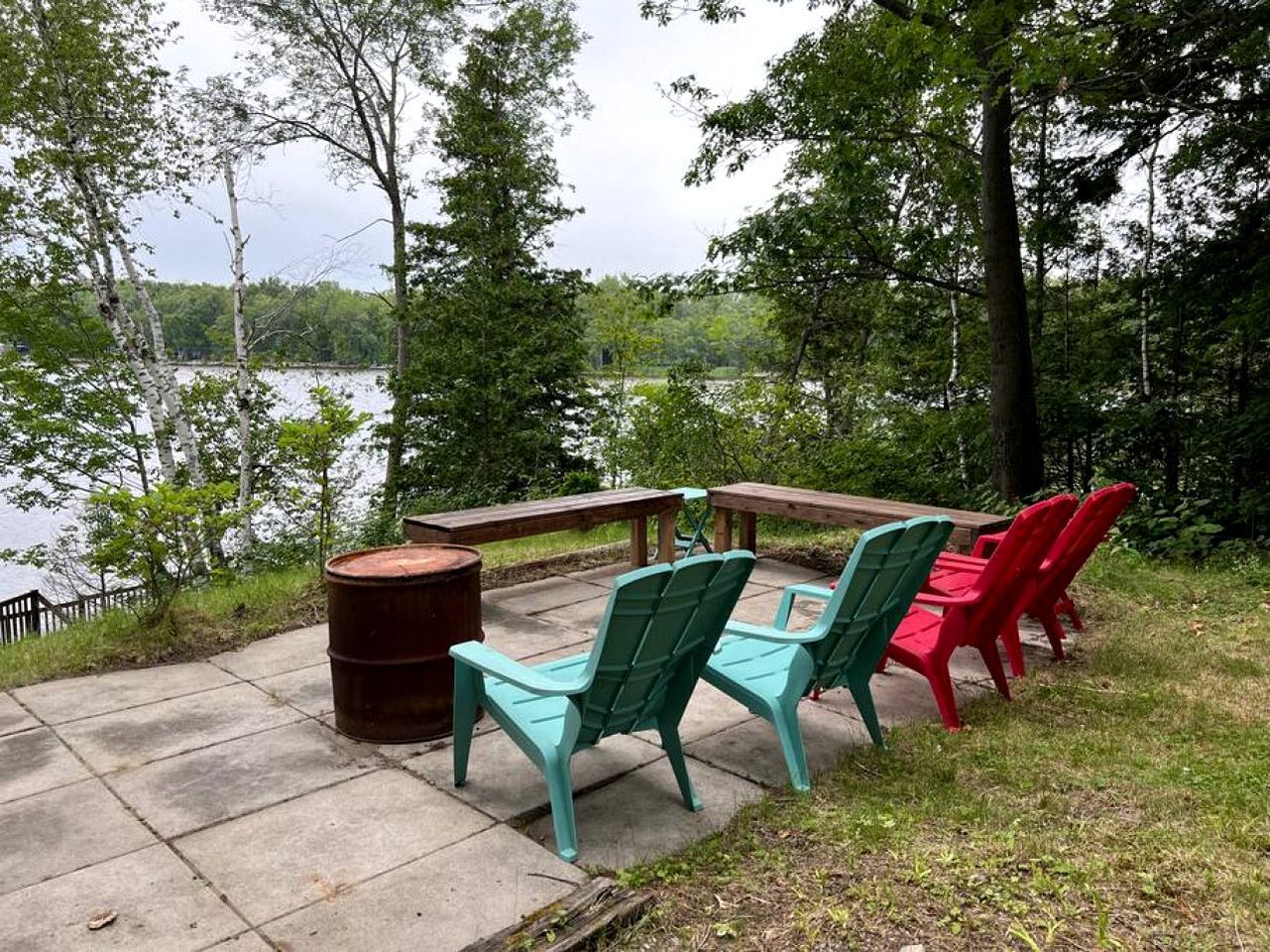 Un patio junto al lago con sillas Adirondack, una mesa de madera y un barril.