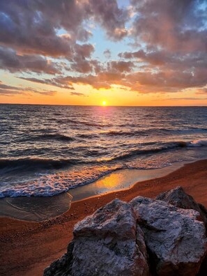 Uma praia com ondas, um pôr do sol e pedras em primeiro plano.