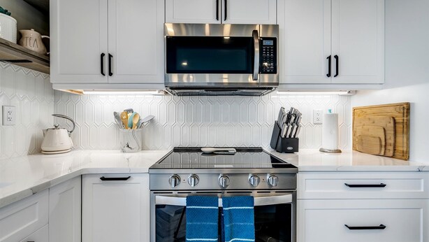 A kitchen with white cabinets, a stainless steel microwave, a gas stove, and a wooden cutting board. - Coast Rental with Hot-Tub and Equipped Deck in Ontario (Leamington)