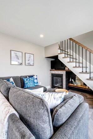 A modern living room with a grey sofa, a fireplace, and a staircase. - Coast Rental with Hot-Tub and Equipped Deck in Ontario (Leamington)