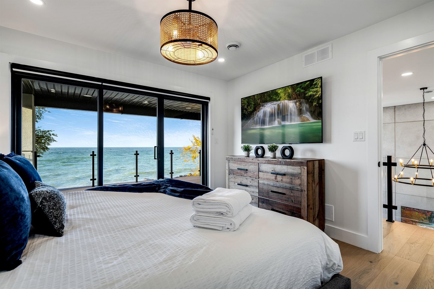 A bedroom with a large bed, a view of the sea, a wooden dresser, and a hanging light fixture.
