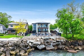 A modern two-story house with a patio and a stone retaining wall. - Sophisticated Shoreline Retreat with Sunset Patio & Poolside Lounging in Ontario, Canada (Leamington)