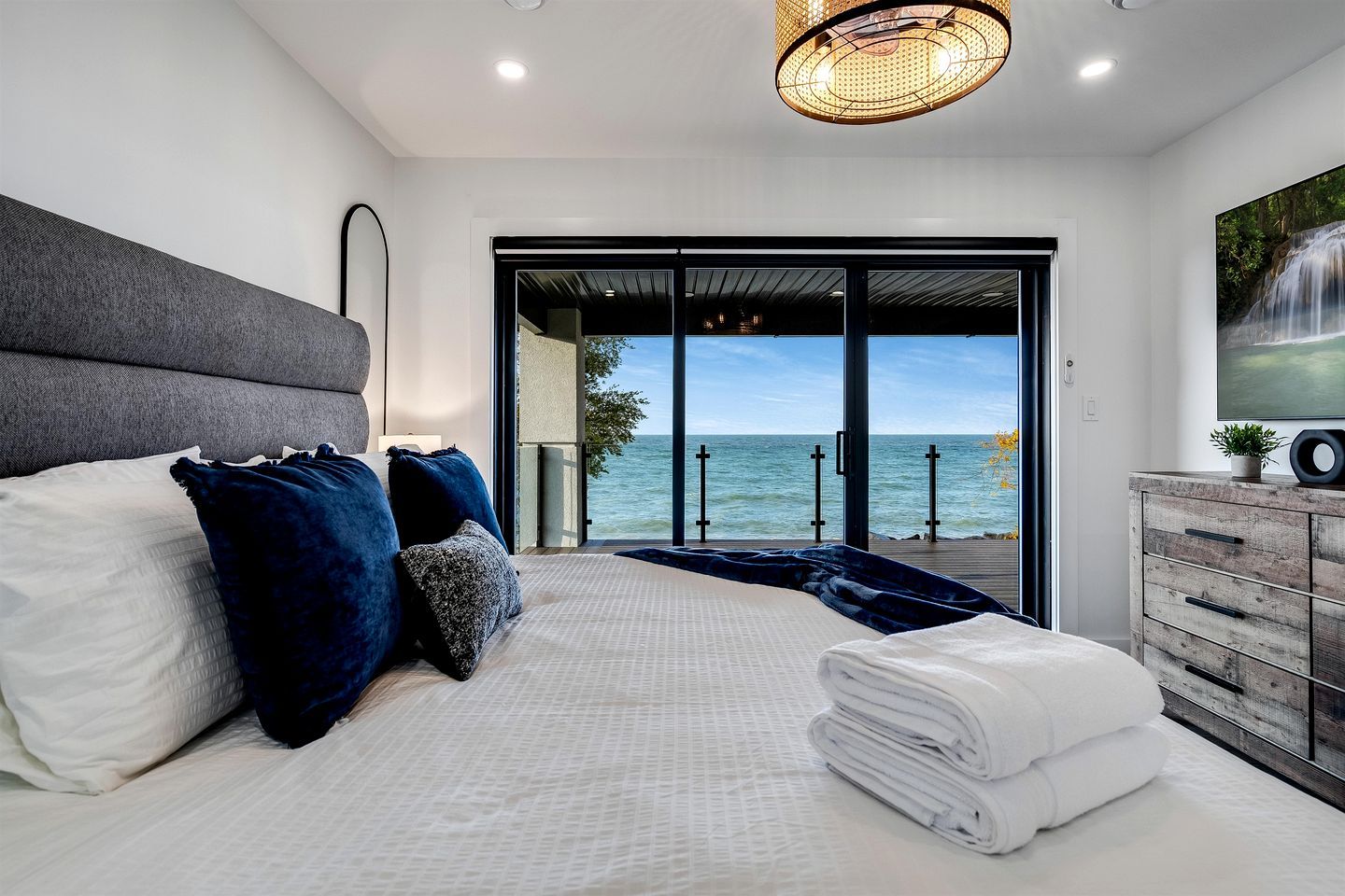 A bedroom with a bed, pillows, a dresser, and a view of the sea.