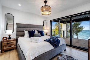 A modern bedroom with a large bed, a wooden nightstand, a mirror, and a view of the sea through large windows. - Sophisticated Shoreline Retreat with Sunset Patio & Poolside Lounging in Ontario, Canada (Leamington)