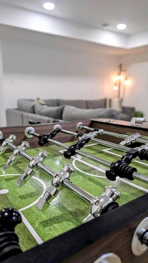 Une table de baby-foot dans une pièce avec un canapé gris et une lampe en arrière-plan.