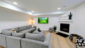 A modern living room with a grey sectional sofa, a flat-screen TV, a fireplace, and a 'Welcome Home' sign. - Sophisticated Shoreline Retreat with Sunset Patio & Poolside Lounging in Ontario, Canada (Leamington)