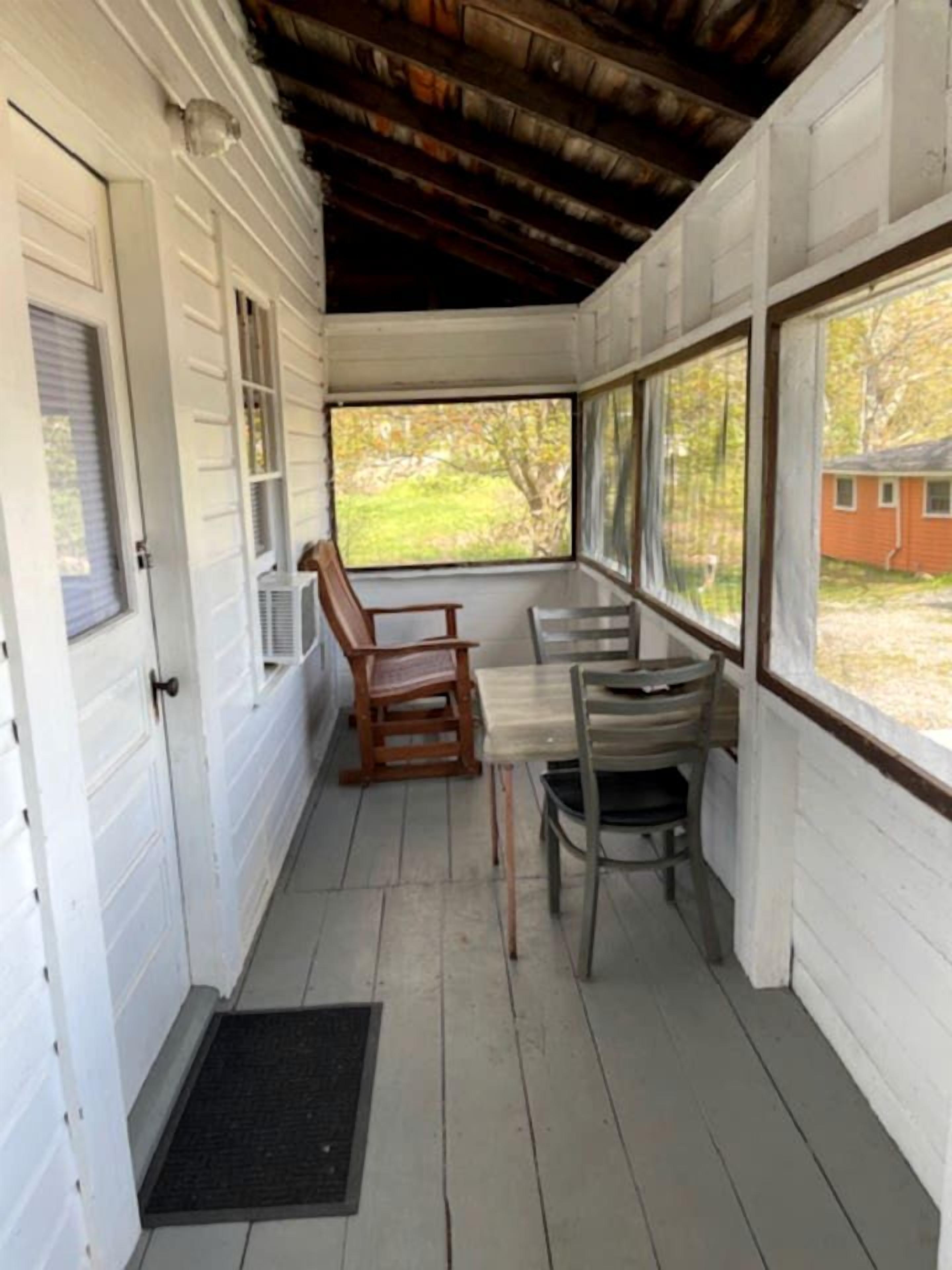 A covered porch with a wooden table and chairs, a door, and a view of a house.