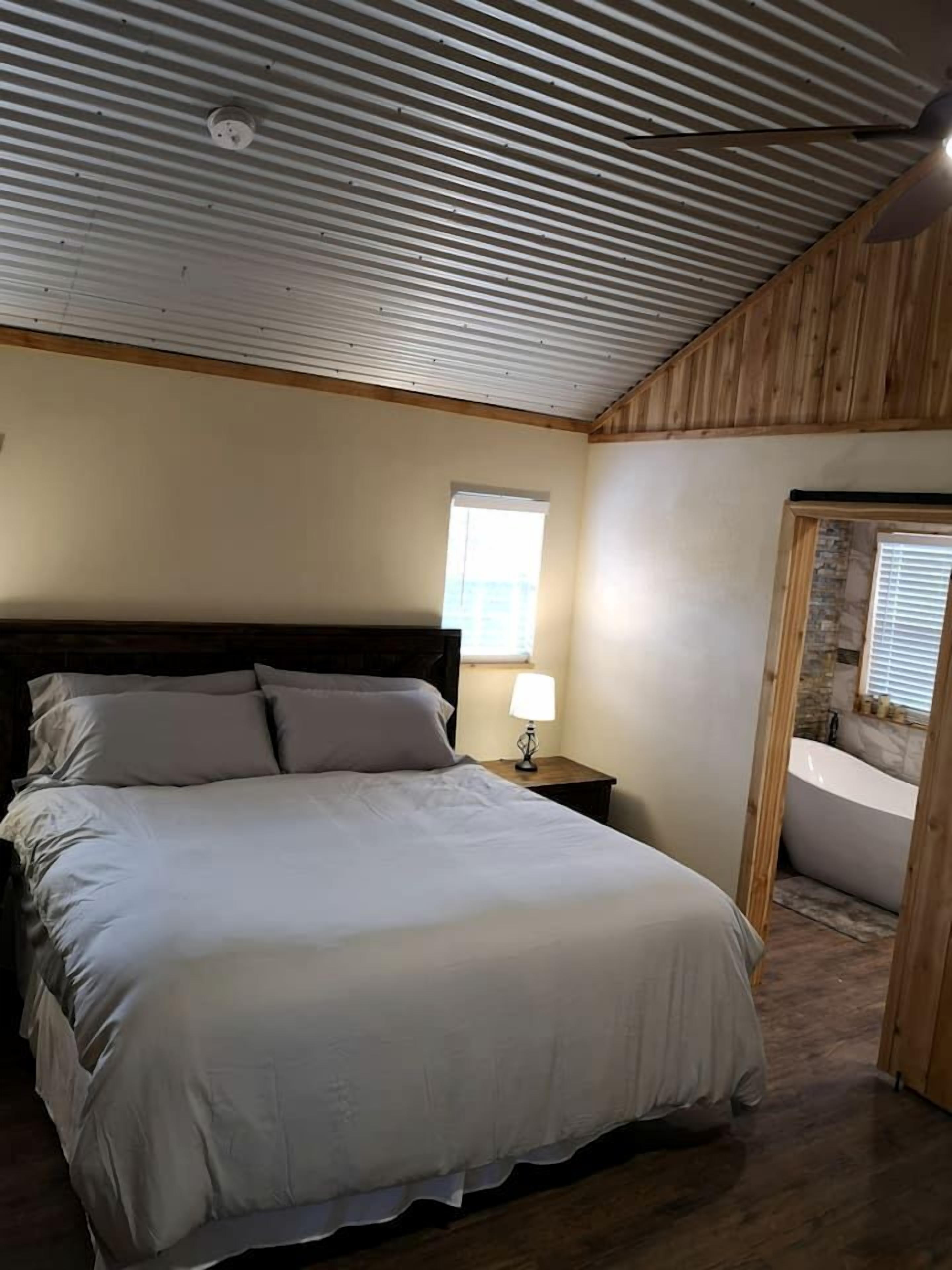 A bedroom with a bed, a nightstand, a window, and a bathroom with a bathtub.