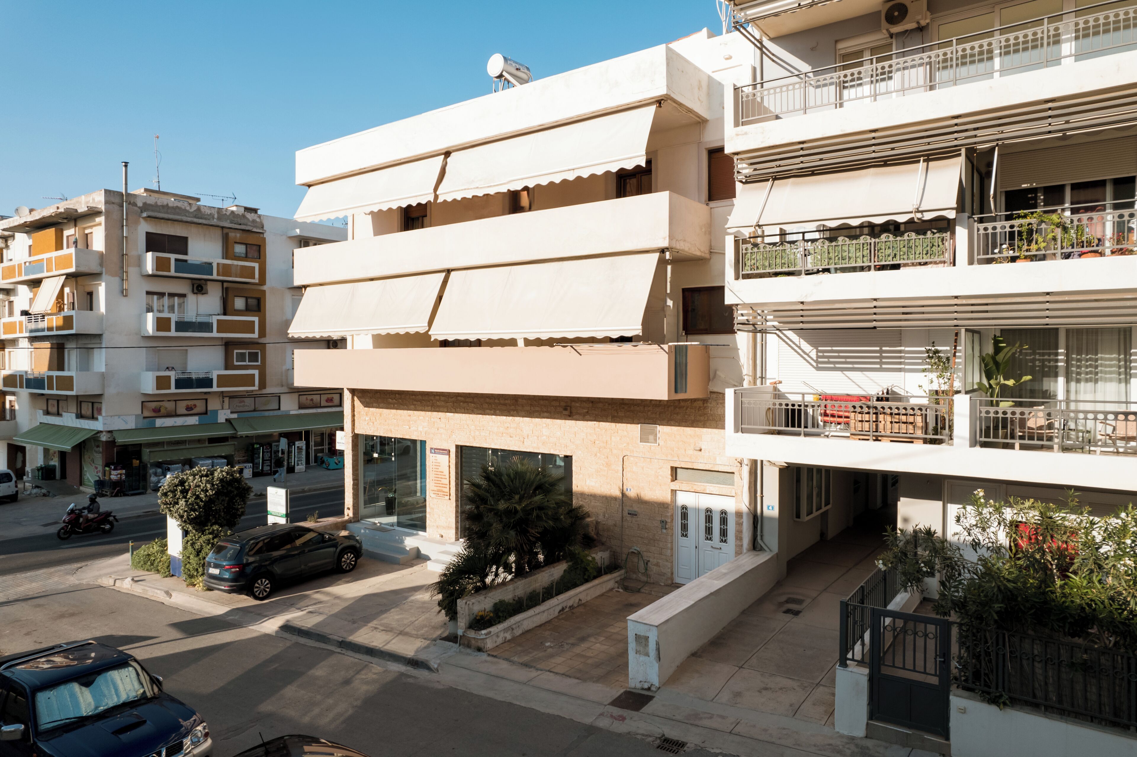 Un edificio residencial de varios pisos con balcones, un coche estacionado adelante y cielo despejado.
